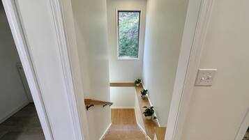 Un couloir étroit avec un escalier en bois, des murs blancs et une petite fenêtre donnant sur la verdure.