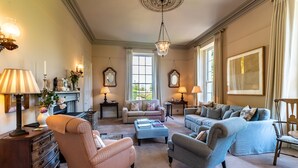 A spacious living room with a fireplace, multiple seating options including a sofa and armchairs, and a chandelier. - Luxury Country Estate in West Cork (Bandon)