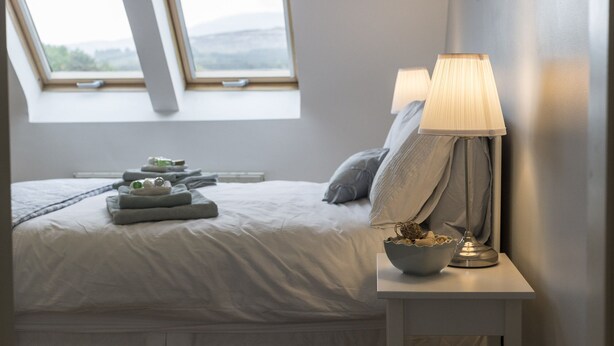 Un lit bien fait, avec une couette blanche, une table de chevet avec une lampe et un bol de fruits.