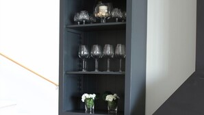 Un meuble haut, étroit et de couleur sombre, doté de plusieurs étagères, dont certaines contiennent de la verrerie et des fleurs.