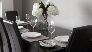 Un coin repas avec une longue table dressée pour six personnes, agrémenté de chaises noires, d’assiettes blanches et d’un vase contenant des fleurs blanches.