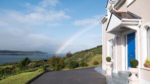 Ein Haus an der Küste mit blauer Tür, befestigter Auffahrt und Blick auf das Meer mit einem Regenbogen.