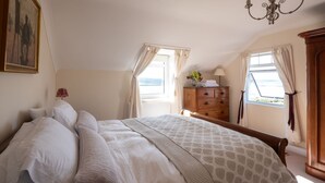 Ein Schlafzimmer mit einem großen Bett, einer hölzernen Kommode, einem Bild an der Wand und Blick durch das Fenster.