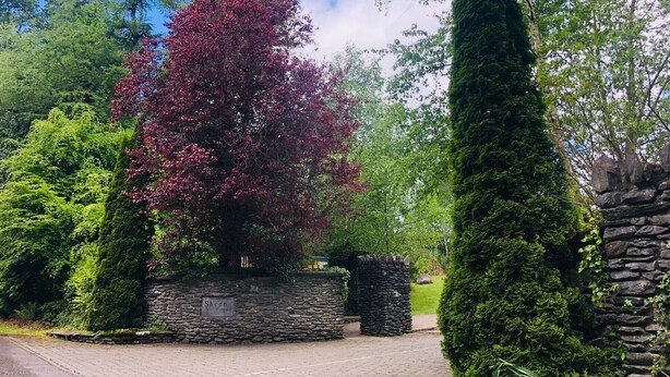 A stone wall with a sign reading "Sheen Woods" and a pathway leading through a lush green landscape.