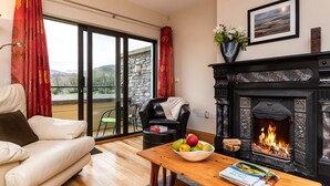 Una sala de estar con chimenea, un sofá, una mesa de comedor con un frutero y libros, y vista a un edificio de piedra a través de un vidrio.