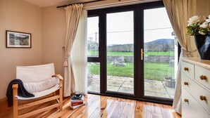 Una habitación con cama, una silla de madera, un aparador y vistas a un paisaje verde a través de un ventanal grande.