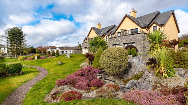Una casa grande y moderna con fachada de piedra y varias chimeneas, rodeada de jardines cuidados y un camino sinuoso.