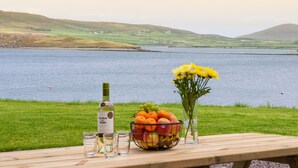 A table with a bottle, a fruit bowl, and a vase of flowers overlooking a lake with hills in the background. - Luxury 4 bedroom holiday home overlooking the sea on Valentia Island (Farranreagh)
