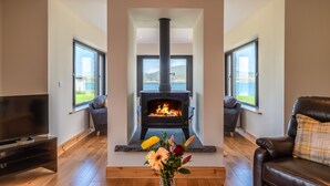 A modern living room with a central wood-burning stove, large windows, a flat-screen TV, and a comfortable seating area. - Luxury 4 bedroom holiday home overlooking the sea on Valentia Island (Farranreagh)