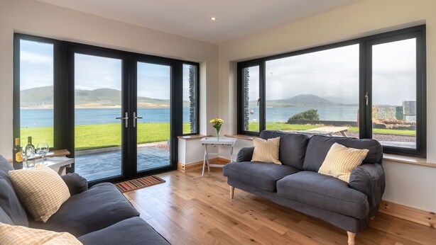A modern living room with a grey sofa, a small table with a vase of flowers, and large windows offering a view of the sea and hills. - Luxury 4 bedroom holiday home overlooking the sea on Valentia Island (Farranreagh)