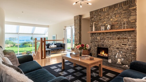 A spacious living room with a stone fireplace, a wooden coffee table, and a view of a lush garden through large windows. - Stylish 4 bedroom home overlooking Kenmare Bay (Templenoe)