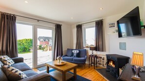 A cozy living room with a sofa, a blue armchair, a wooden coffee table, a fireplace, and a TV mounted on the wall. - Large house with adjoining cottage close to Waterville (Waterville)