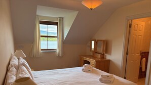 A bedroom with a large bed, a nightstand, a dresser, a mirror, and a window with curtains. - Holiday Home with view of Kenmare Bay Estuary (Kenmare)