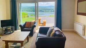 Ein Wohnzimmer mit Fernseher, einer Couch, einem Holztisch und Blick auf einen See und Berge durch große Fenster.