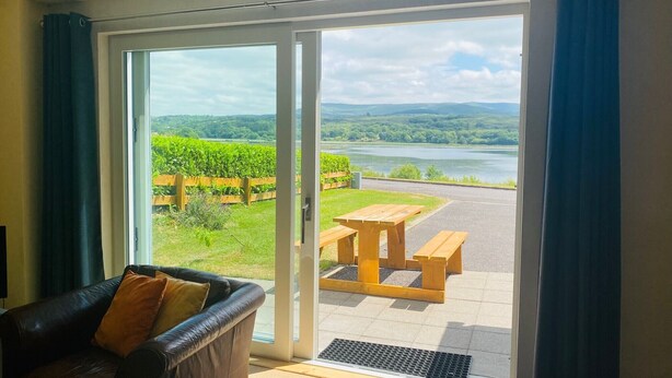 Ein Zimmer mit einer Schiebeglastür, die zu einem Außenbereich mit Picknicktisch und Bänken führt. Blick auf einen Fluss und eine Rasenfläche mit Holzzaun.