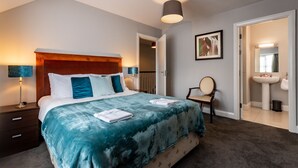A bedroom with a large bed, a bedside table, a lamp, a chair, a painting, and a bathroom visible through an open door.