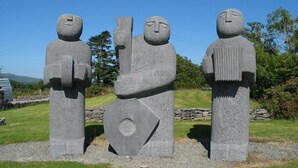 Un monument de pierre représentant trois personnages, chacun tenant un bâton, est érigé dans un espace verdoyant, agrémenté d’arbres, avec un ciel dégagé en arrière-plan.