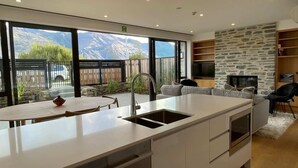 A modern kitchen with a large island, a dining area with a table and chairs, a stone accent wall, and a view of mountains through large windows. - Central Wanaka Lakeside Condo (Wanaka)