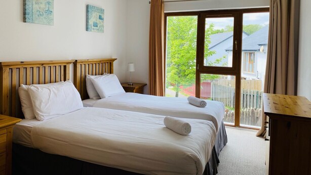 A hotel room with two beds, a wooden headboard, a nightstand with a lamp, and a view of the outside. - Modern home with 3 bedrooms and 3 bathrooms in Kenmare town (Kenmare)