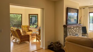 Una sala de estar con muebles de mimbre, una chimenea de piedra y grandes ventanales con vistas a la vegetación.
