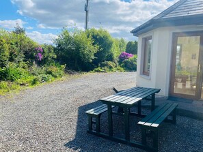 A gravel area with a wooden picnic table and benches, surrounded by lush greenery and purple flowers.