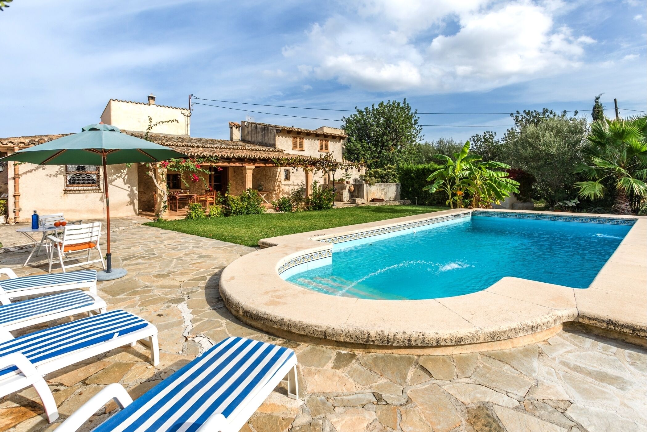 A swimming pool with stone tiles, a patio with lounge chairs, and a building with a tiled roof.