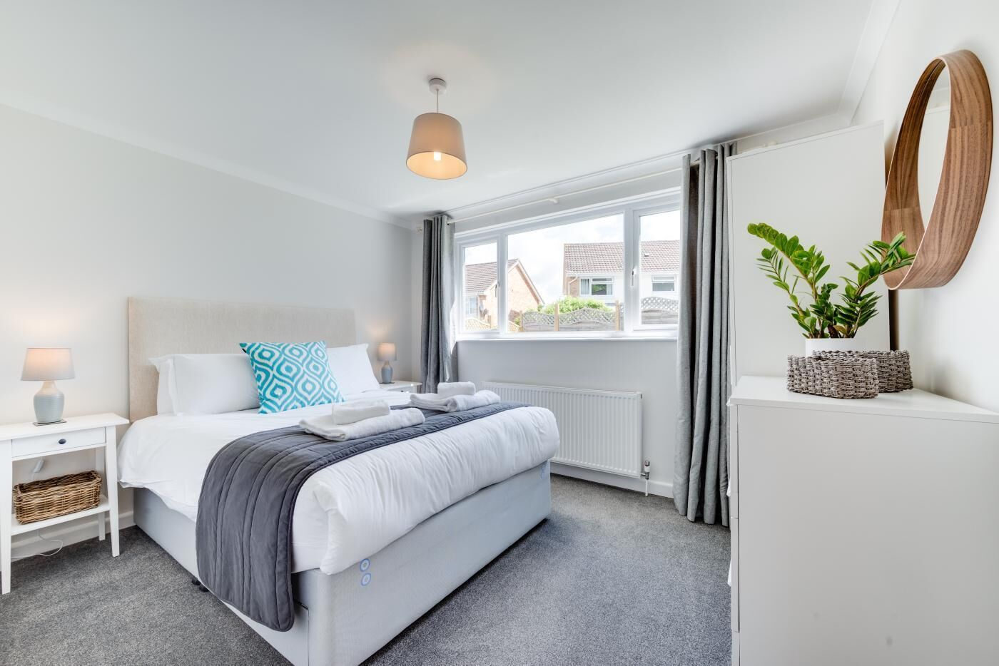 A modern hotel bedroom with a bed, bedside tables, a radiator, a window with curtains, and a potted plant.