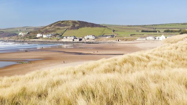 Un paysage côtier avec une plage de sable, des dunes herbeuses et une colline sur laquelle se trouvent des bâtiments en arrière-plan.