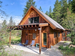 A wooden cabin with a porch, two chairs, a bench, and a gravel path.
