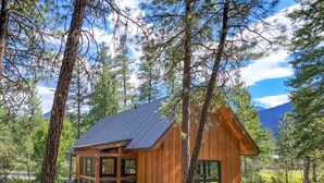 A wooden cabin with a sloped roof, large windows, and a porch area, surrounded by trees and a mountainous backdrop. - Adeline - cute Mazama meadow cabin with sauna (Mazama)