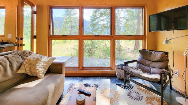 A cozy room with a large window, a sofa, a chair, a TV, and a remote control on a wooden table. - Adeline - cute Mazama meadow cabin with sauna (Mazama)
