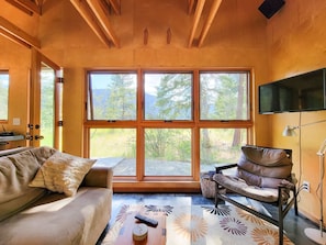 A cozy room with a large window, a sofa, a chair, a TV, and a remote control on a wooden table.