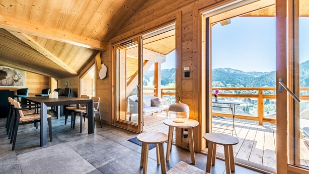 Una cabana de fusta amb zona de menjador, un balcó amb vistes a les muntanyes i un dormitori acollidor amb un llit i una nitzeta.