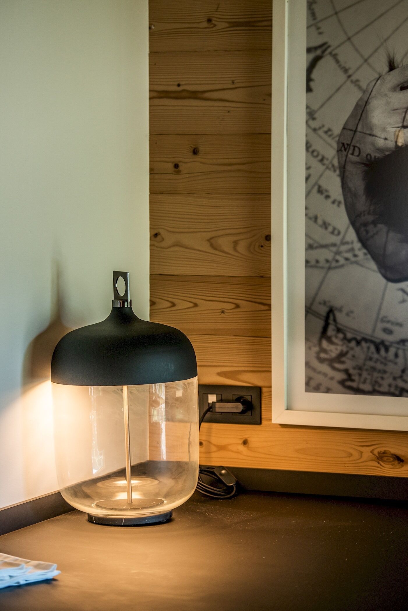 Uma luminária de mesa com cúpula preta e base transparente, sobre uma superfície de madeira ao lado de um mapa emoldurado.