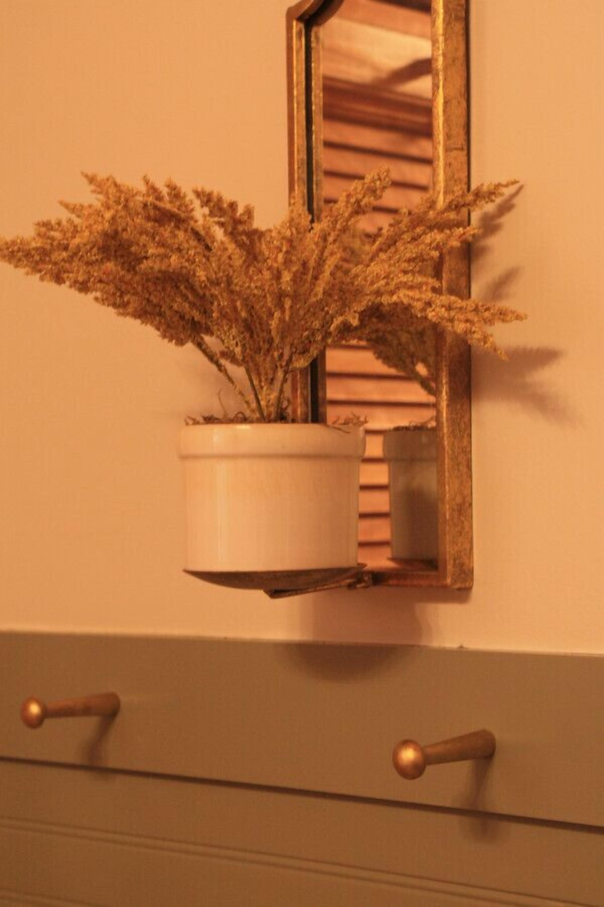 Una planta en maceta con flores secas sobre un estante fijado a la pared, encima de un mueble con dos cajones.