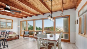 A spacious room with a wooden ceiling, large windows, a dining table with chairs, and a view of the outdoors.
