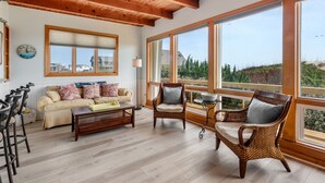 A spacious living room with a wooden ceiling, large windows, a sofa, two wicker chairs, a coffee table, and a clock on the wall.