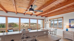 A modern kitchen with a large island, a ceiling fan, and large windows offering a view of the beach.
