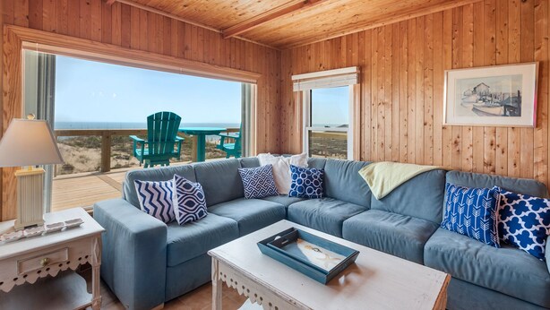 A wooden-paneled room with a blue sectional sofa, a small white table, and a view of the beach.