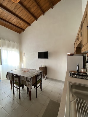 A kitchen with a dining table, chairs, a TV mounted on the wall, and a refrigerator.