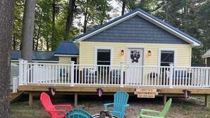 A yellow vacation rental with a white deck, blue and red Adirondack chairs, and a sign indicating the rental name and contact number.