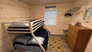 A compact cabin room with a bunk bed, a wooden dresser, a small rug, and a window with blinds.