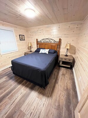 Un dormitorio con cabecera de madera, cama con colcha oscura, dos mesitas de noche con lámparas y una ventana con persianas.