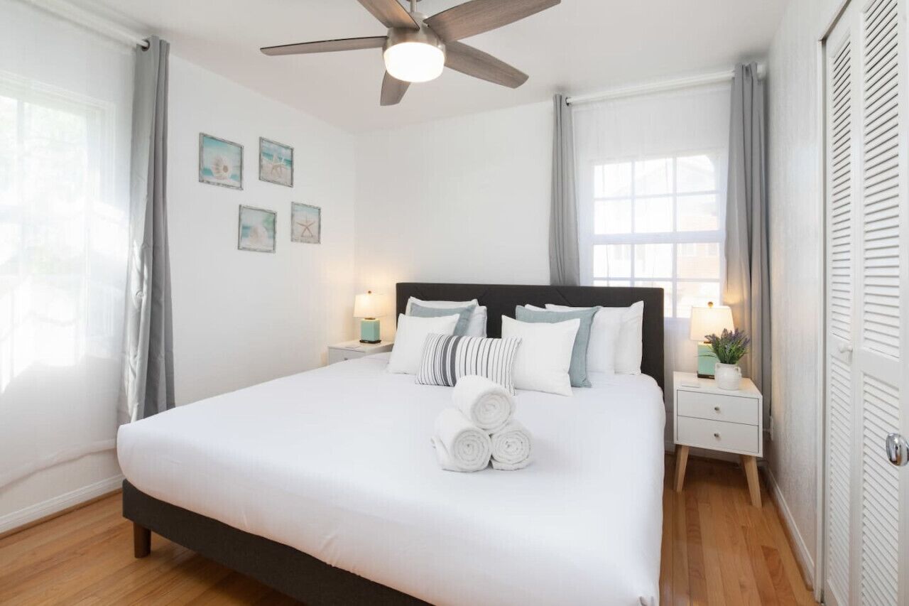 Una cama bien tendida con ropa de cama blanca, un ventilador de techo, mesitas de noche con lámparas y una ventana con cortinas.
