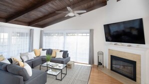 A modern living room with a grey sofa set, a glass coffee table, a fireplace, and a large TV mounted on the wall.
