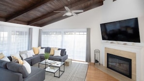 A modern living room with a grey sofa set, a glass coffee table, a fireplace, and a large TV mounted on the wall.