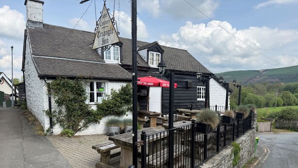 Un bâtiment traditionnel avec un toit triangulaire, une enseigne indiquant « Auberge du Triangle, XVIe siècle », et des tables et des bancs à l’extérieur pour les clients.