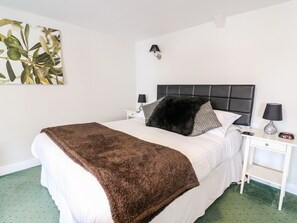 A neatly made bed with a fluffy brown blanket, a black and white striped pillow, and a black fluffy pillow. There is a bedside table with a lamp and a clock. The room has white walls and a green carpet. - Triangle Inn Coachouse (Rhayader)