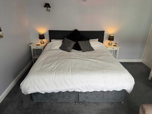 A neatly made bed with white bedding and two decorative pillows, flanked by bedside tables with lamps, in a room with a dark headboard and light walls.