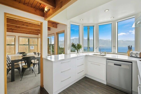 A modern kitchen with white cabinetry, a wooden ceiling, and large windows offering a mountain view.